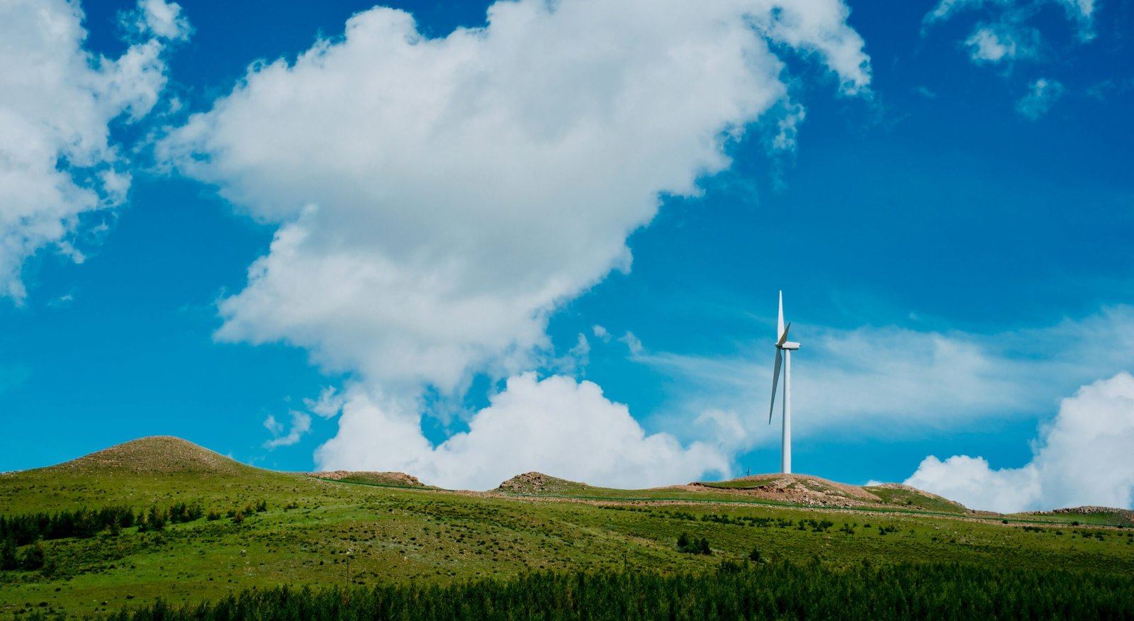 Wind Turbine Power Generation in Mountain Landscape A wind turbine standing on a green hillside under a bright blue sky with clouds, generating clean and renewable wind energy.
